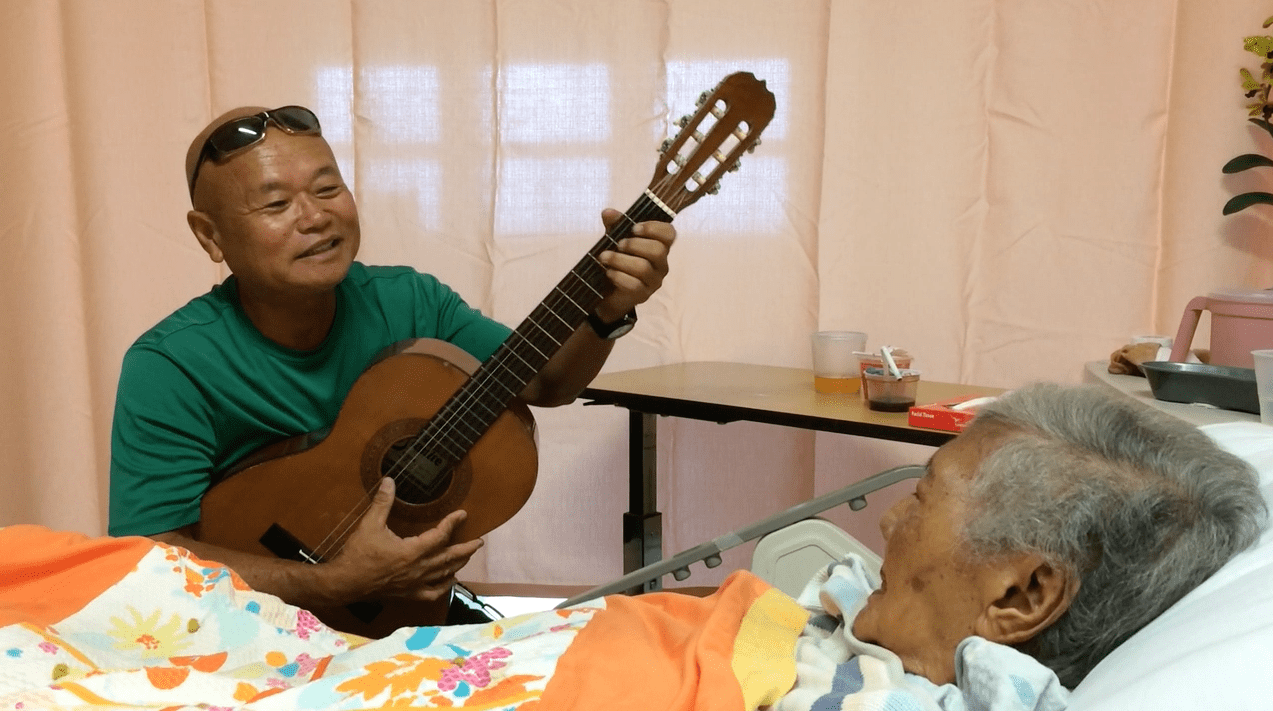 A man plays guitar beside a bedridden elderly person in a cozy room.
