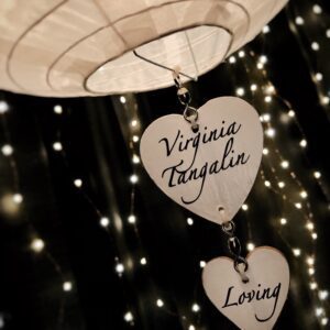 Heart-shaped pendants with 'Virginia Tagalio' and 'Loving' engraved, hanging against blurred lights.